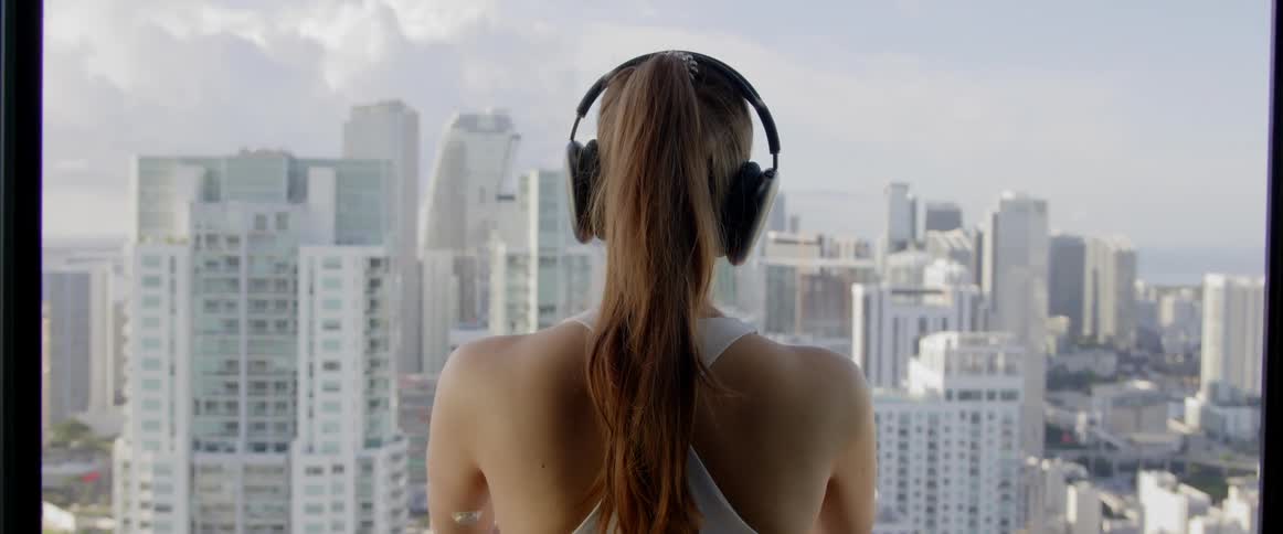 Back view of a woman with headphones looking out a window overlooking a metropolis
