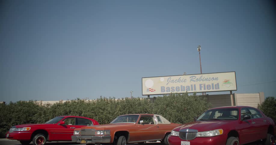 Three cars sit in a parking lot next to a sign for a baseball field