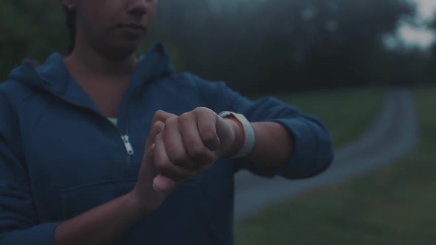 A woman in a hoodie looks at her smart watch while standing by a small path