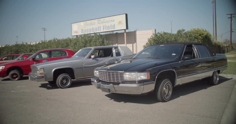 Four lowrider cars parked in a parking lot in front of a baseball field