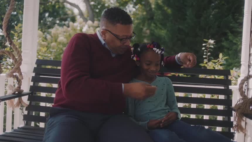 Man in red sweater with his daughter girl on a bench in an outdoor setting with trees and greenery in the background