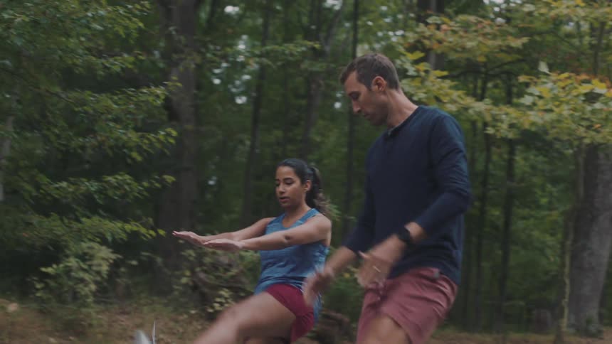 While on a road near the woods a male trainer guides a woman in an exercise involving high kicks and steps