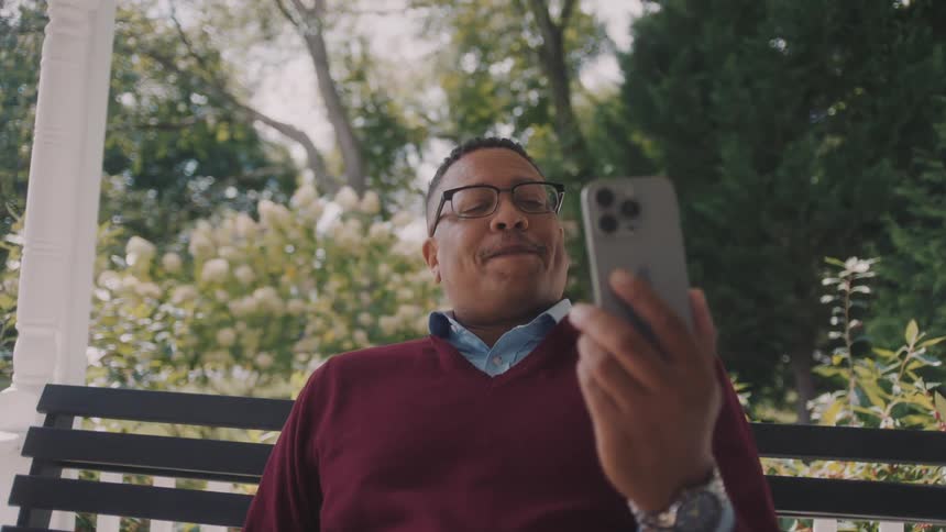 An older black man sits on a porch while talking to someone through a smartphone