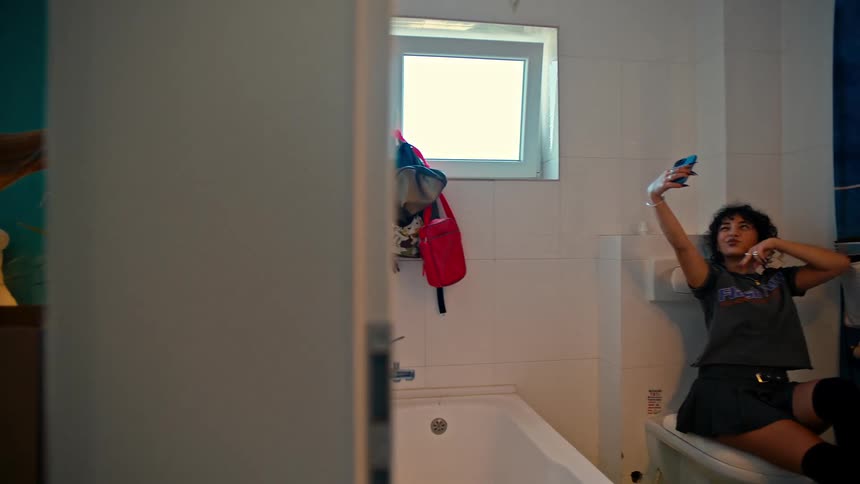 A girl organizes books on a shelf while another takes selfies in the bathroom