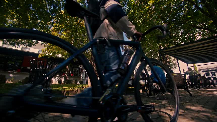 A man carrying a bicycle passes in front of a group of people sitting on a bench in a park working on laptops