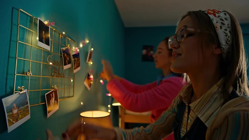 Friends decorate the walls of a dorm room with christmas lights and polaroid photos
