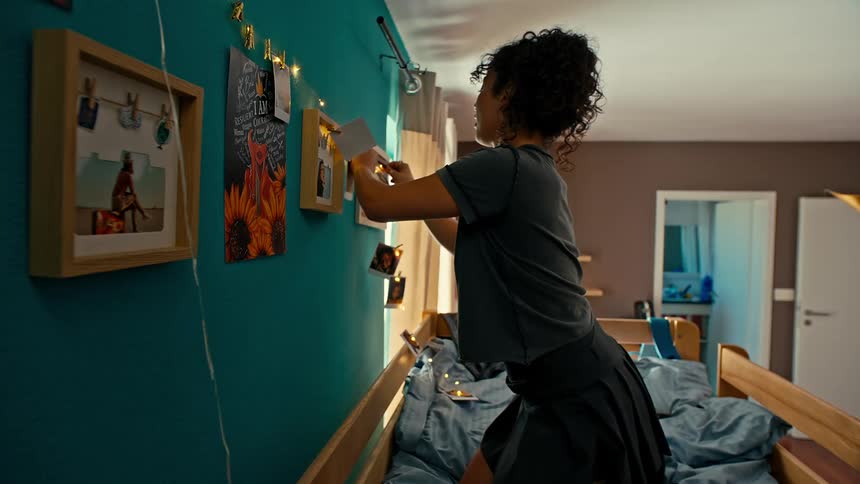 A young girl sits on a bunk bed decorating the bedroom wall with pictures and christmas lights