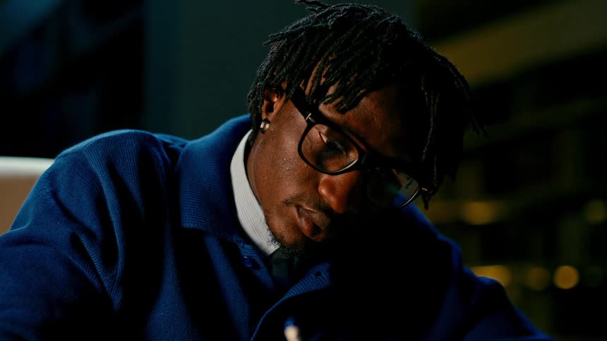 A young man wearing glasses and a blue sweater writes in a notebook and rubs his face in tiredness