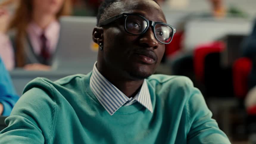 A man sits in a college classroom taking notes and listening to a lecture