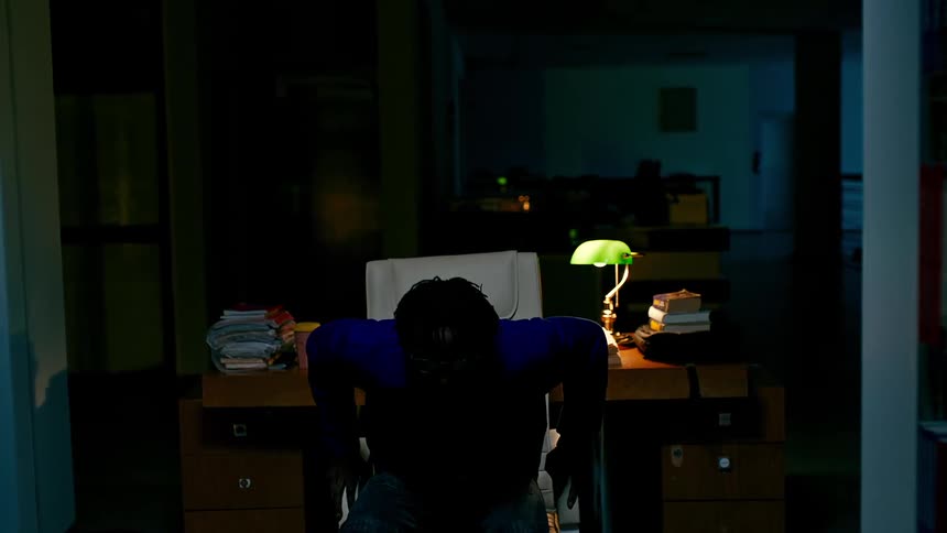 A person rises from a desk and walks down the library aisles at night