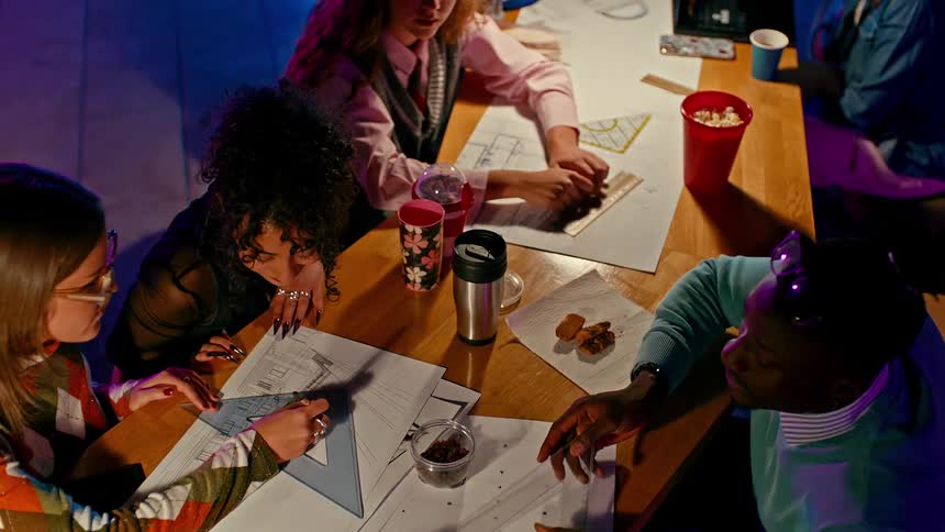 A group of students sit at a large desk studying and drawing blueprints with rulers