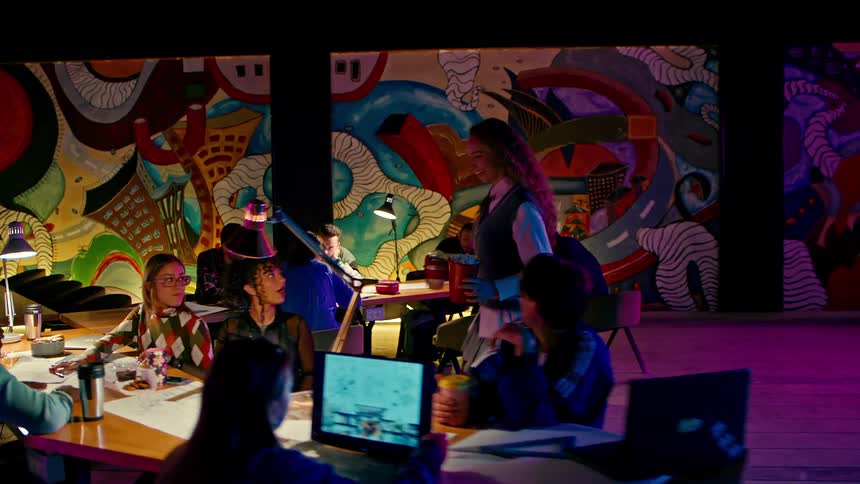 A young woman carries popcorn and a dessert to a table where her friends are laughing and having fun
