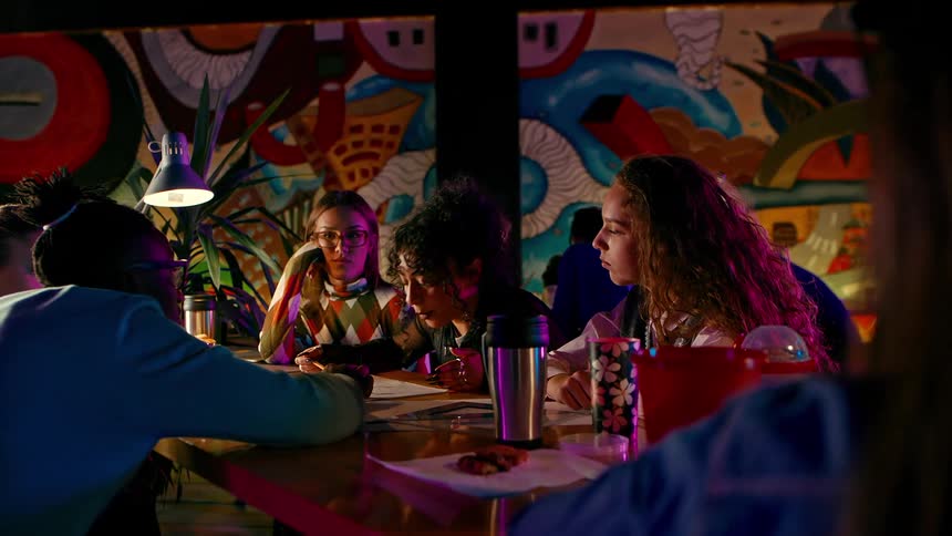A group of friends gathers in a dimly lit colorful cafe to study