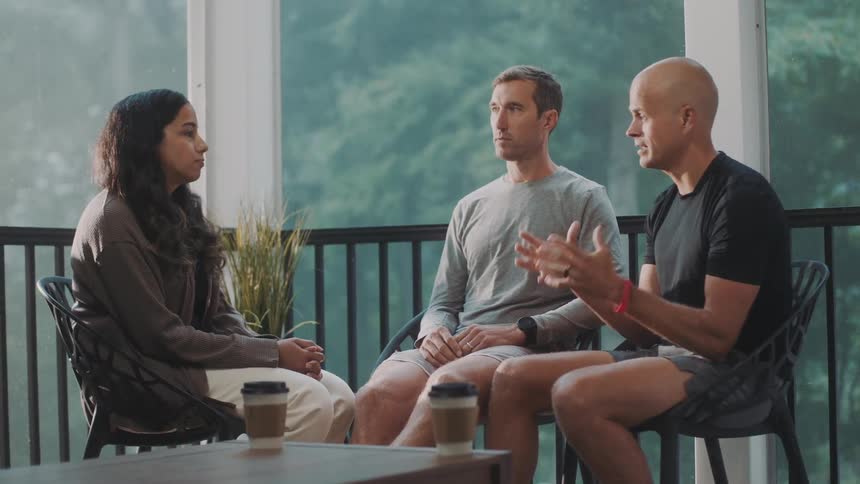 A group of friends sits on a porch or balcony with large windows talking