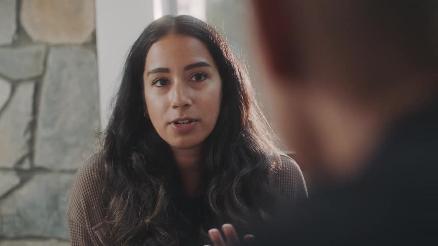 A close up of a woman with long hair talking in a room next to a window
