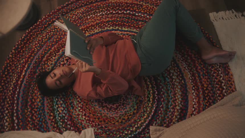 A young woman lying on a rug writes in a notebook