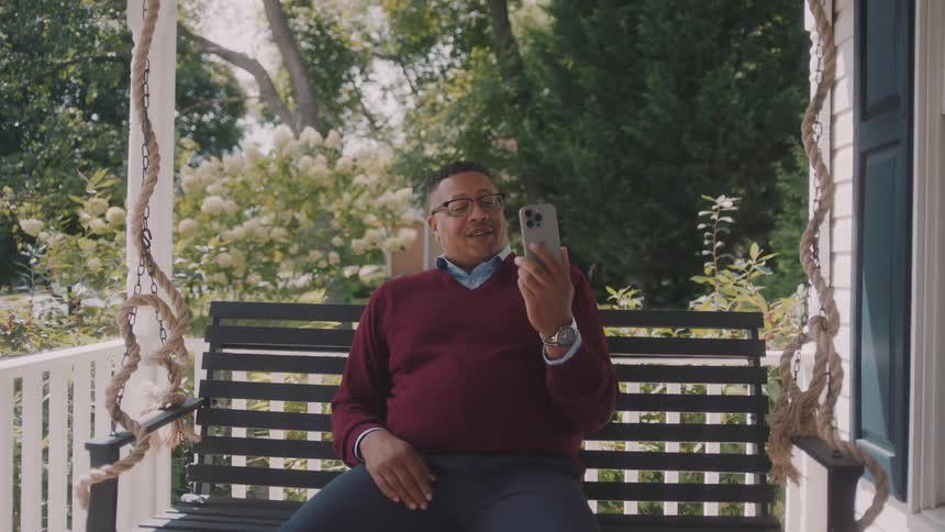 A man sits on a hanging chair on the porch smiling during a video call