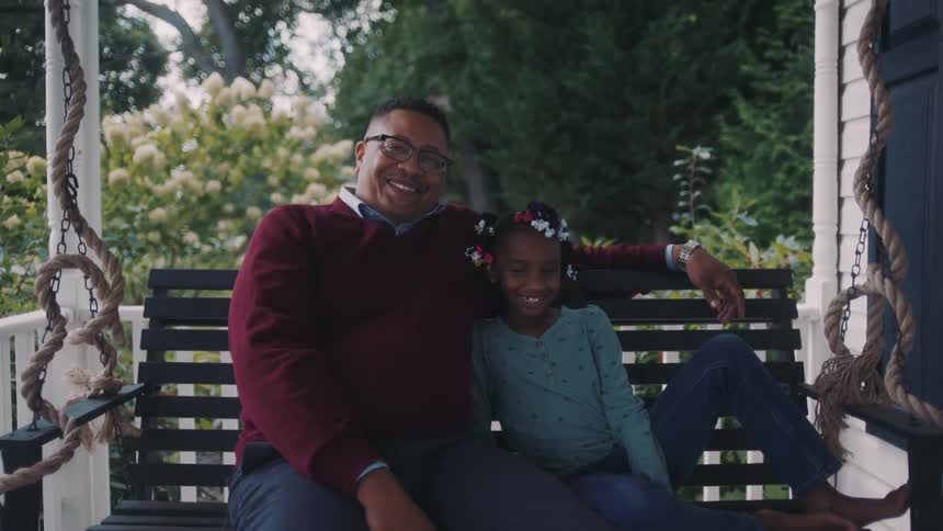 A father and daughter sit on a hanging chair on the porch smiling at the camera