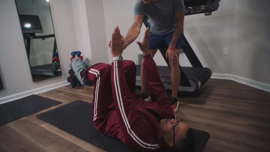 Man standing helping another man who is lying on a yoga mat to exercise his body