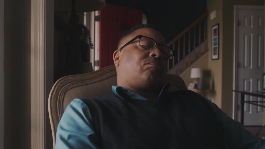 A man wearing glasses is sitting on a chair in a living room and is looking thoughtfully at the window and then looks away