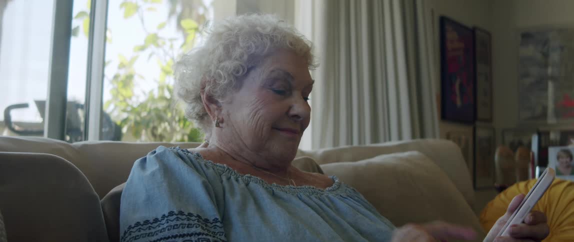 An elderly woman smiles while reading on her smartphone sitting on the couch at home