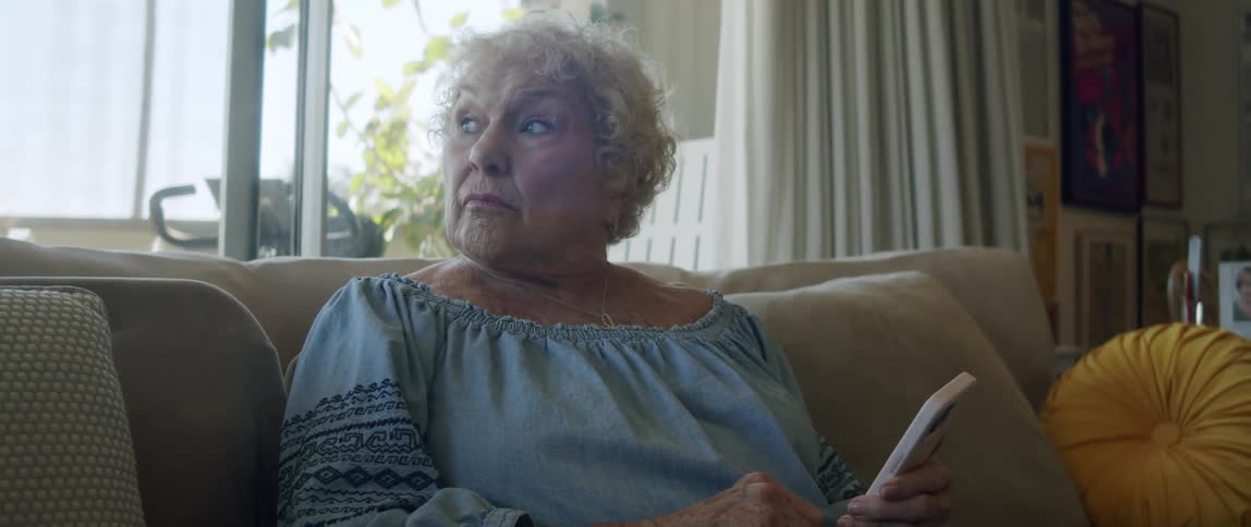 An elderly woman looks out the window while reading on her smartphone sitting on the couch at home