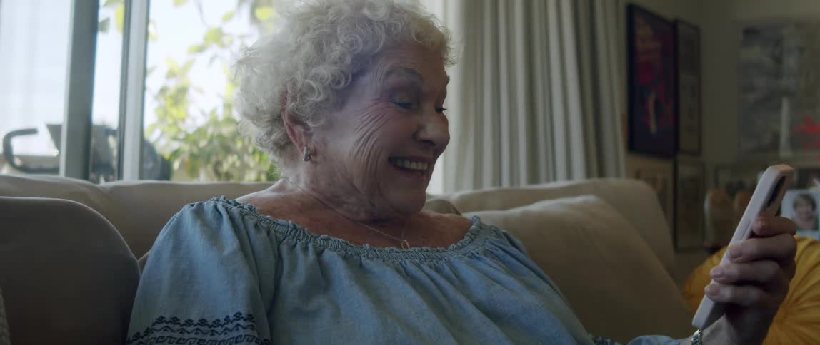 An elderly woman laughs while reading on her smartphone sitting on the couch at home