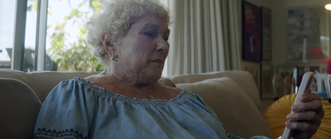 An elderly woman is arguning while reading on her smartphone sitting on the couch at home