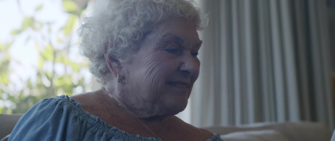 Happy elderly woman sitting in living room next to window and interacting with smartphone