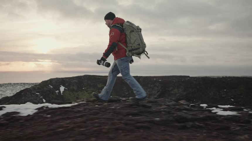 Man with a backpack and a professional camera in his hands climbing a hill in a natural environment surrounded by mountains and the sea