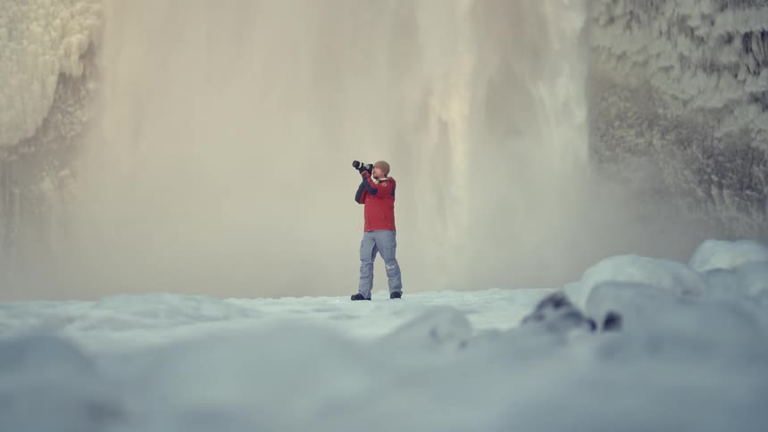 A photographer standing in front of a waterfall on a winter setting taking pictures