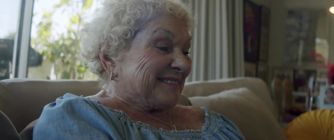An elderly woman laughs while reading on her smartphone sitting on the couch at home