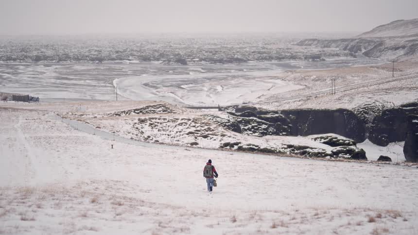 Person walking alone in a snowy landscape with rock formations and a winding road in the background
