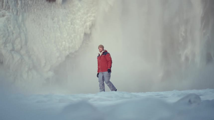 A photographer walks through a snowy landscape carrying his camera toward a waterfall that is partially frozen
