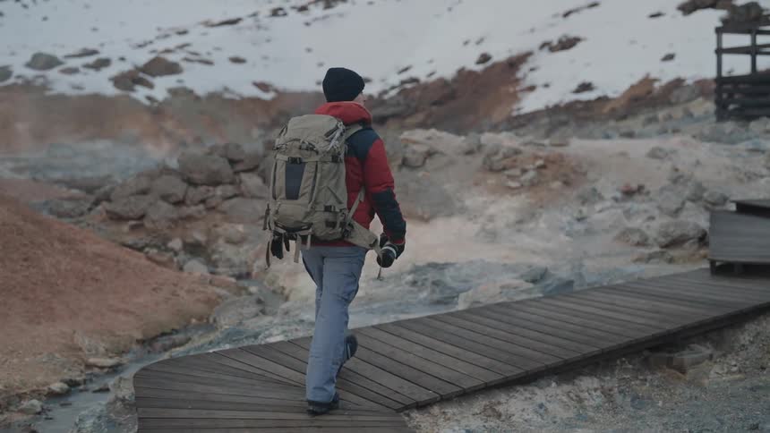 Man walking along a trail in a natural area of snowy mountains with a camera in his hand and a backpack on his back