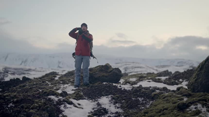 Man with a professional camera using it while standing on rocks with snow and mountains in the background