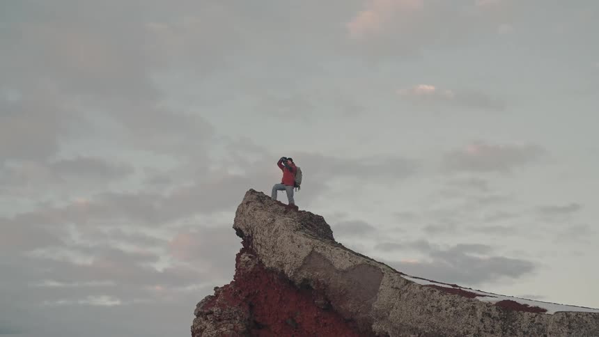 Man with a professional camera standing on the edge of a steep rocky slope against a cloudy sky