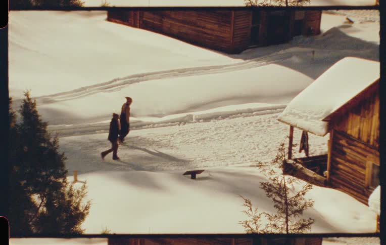 Two people walking through a snowy area with small wooden houses around