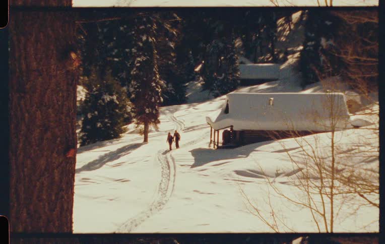 Two people walk along a snowy path next to a cabin in the woods