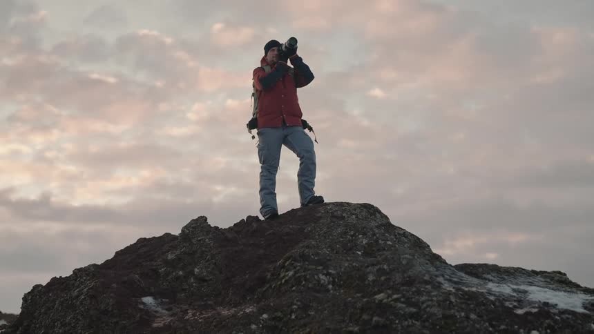 Man standing on top of a rocky outcrop with patches of snow using a professional camera