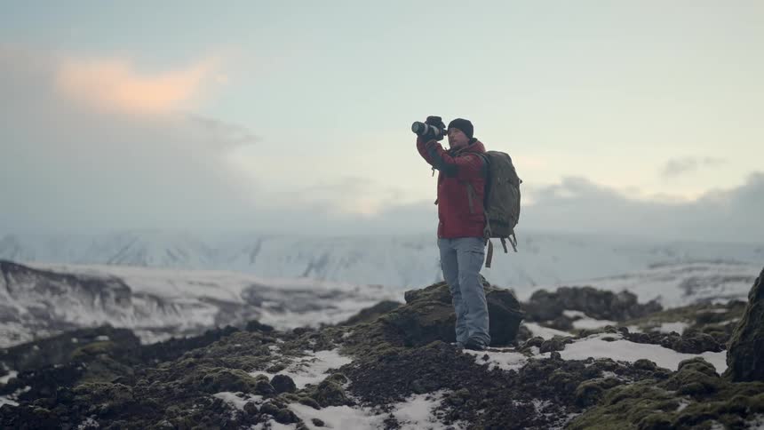 Man on top of rocks in a snowy environment with mountains in the background using a camera with a professional lens