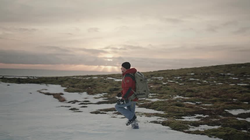 A man with a backpack and warm clothes is walking in a region with the ground covered in snow while he is looking to the sides and carrying a camera in his hand
