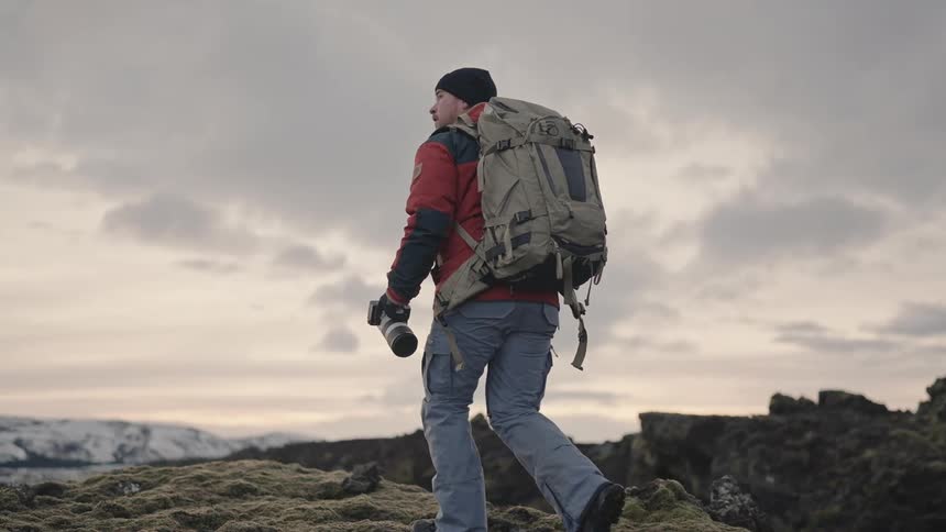 A photographer wearing a red jacket and backpack hikes up a mossy hill and takes a photo