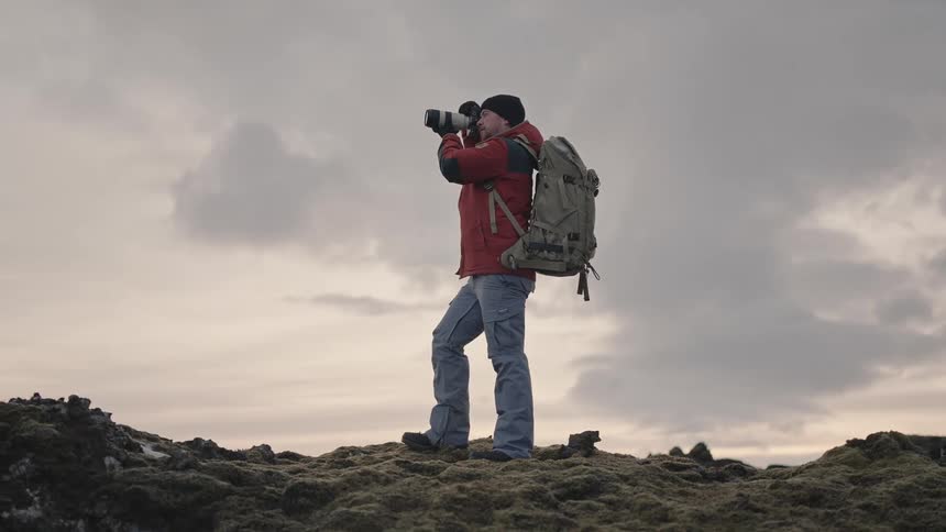 Male photographer using his professional camera while standing on a rock in a snowy natural environment