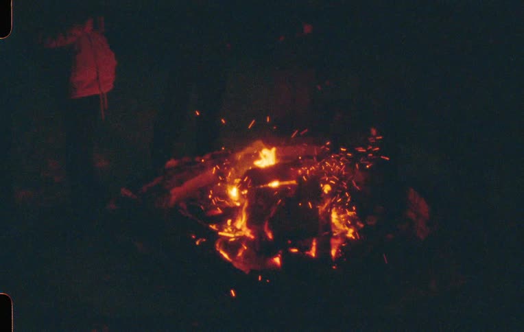 Sparks fly up from a bonfire at night as figures stand around it