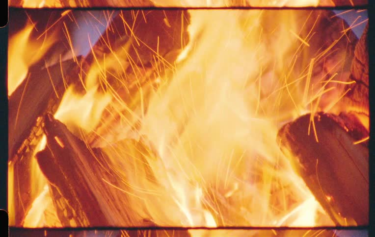 A person moves around behind a roaring campfire outdoors