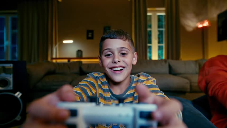 A young boy in a striped shirt is enthusiastically holding a video game controller in front of him with another person smiling beside him on a cozy couch in a warmly lit living room