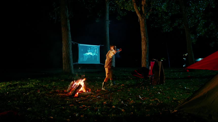 Boy waves a burning stick around a campsite at night stopping occasionally to place it into a burning campfire