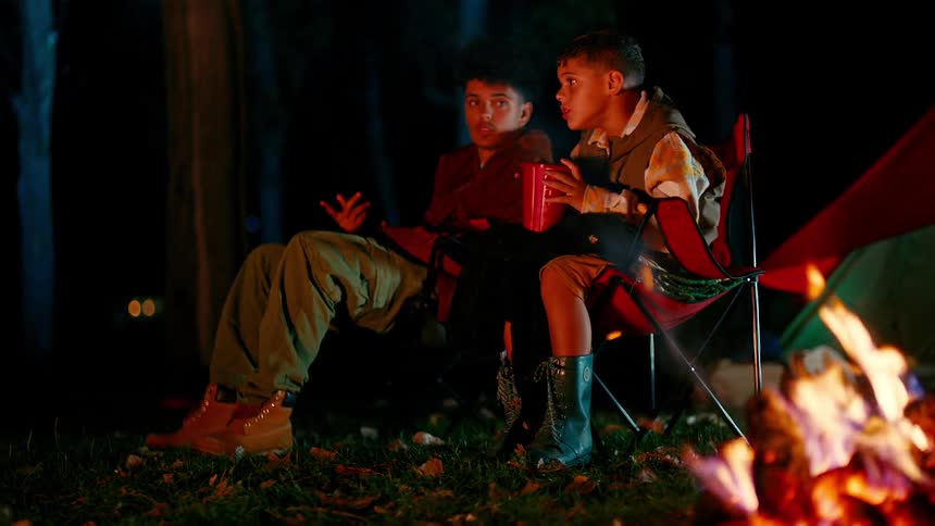 Two boys sitting at a campsite with a campfire burning talking at night