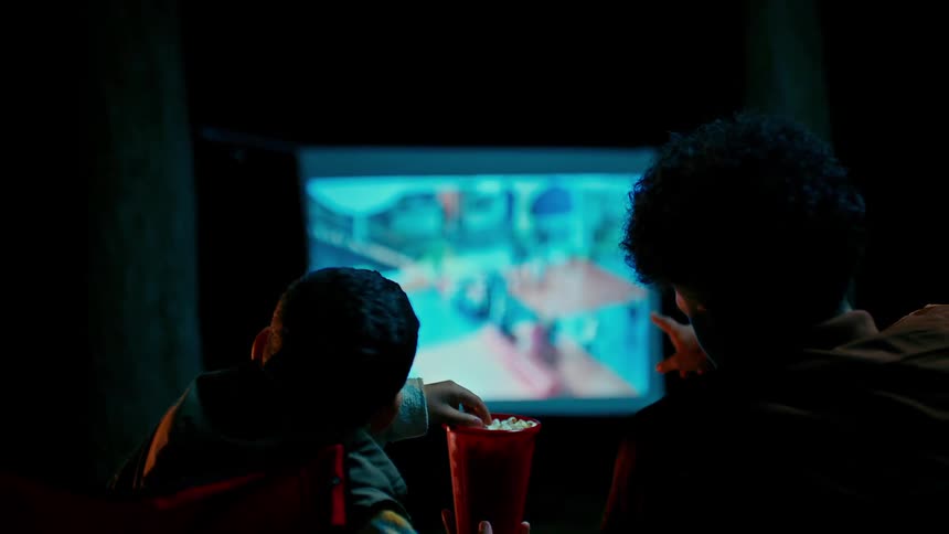 A man and a child sit together in a dimly lit room engrossed in watching a movie on a large screen and eating popcorn